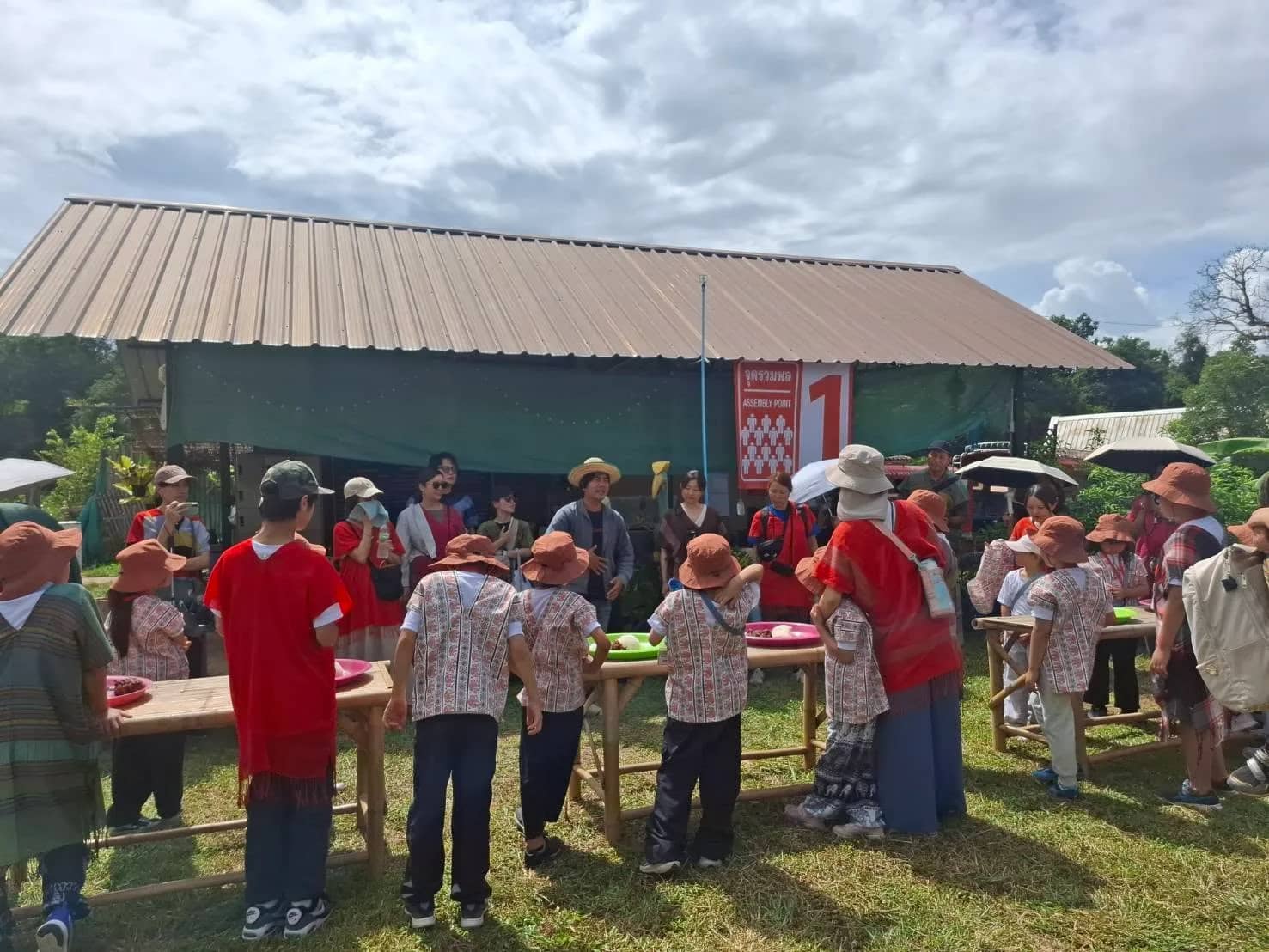 Visitors in Karen shirts and hats gather around tables outside a large farm shelter