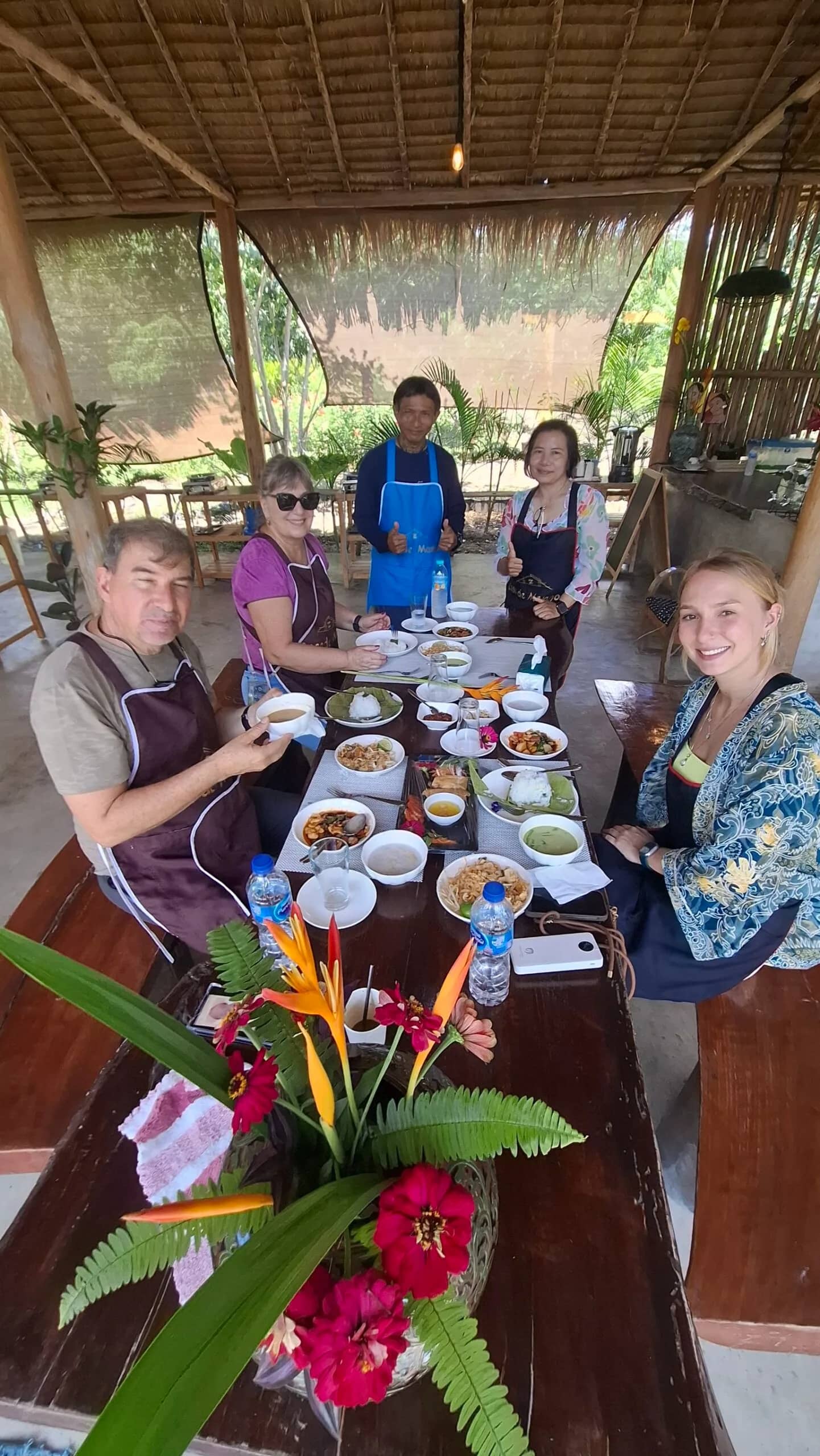 Guests in aprons sit around a long table of Thai dishes in an open-air bamboo kitchen.