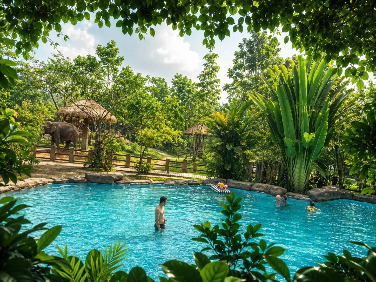A visitor stands in a turquoise pool surrounded by tropical plants with elephants and huts behind
