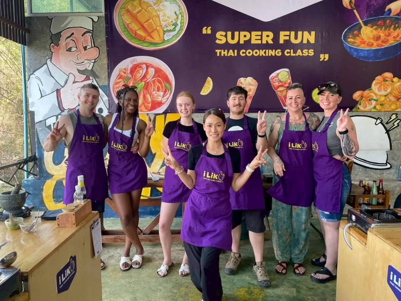 Guests in purple aprons pose in front of a colourful Super Fun Thai Cooking Class mural
