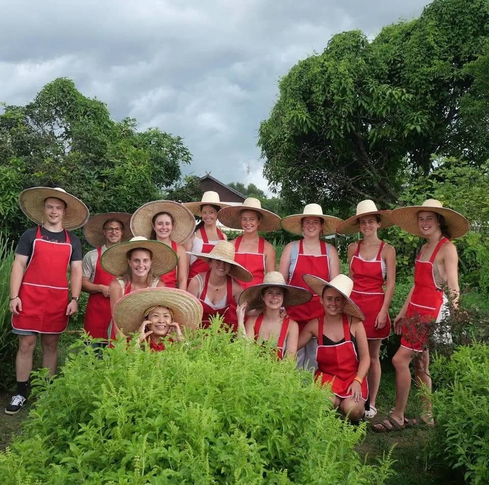 Guests in red aprons and wide straw hats gather among herbs at Smile Organic Farm Cooking School