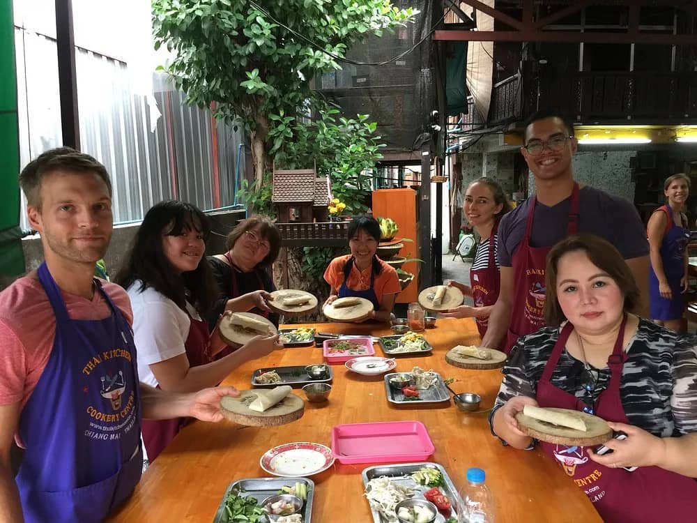 Smiling cooking class guests in purple aprons hold up plates around a long wooden table.