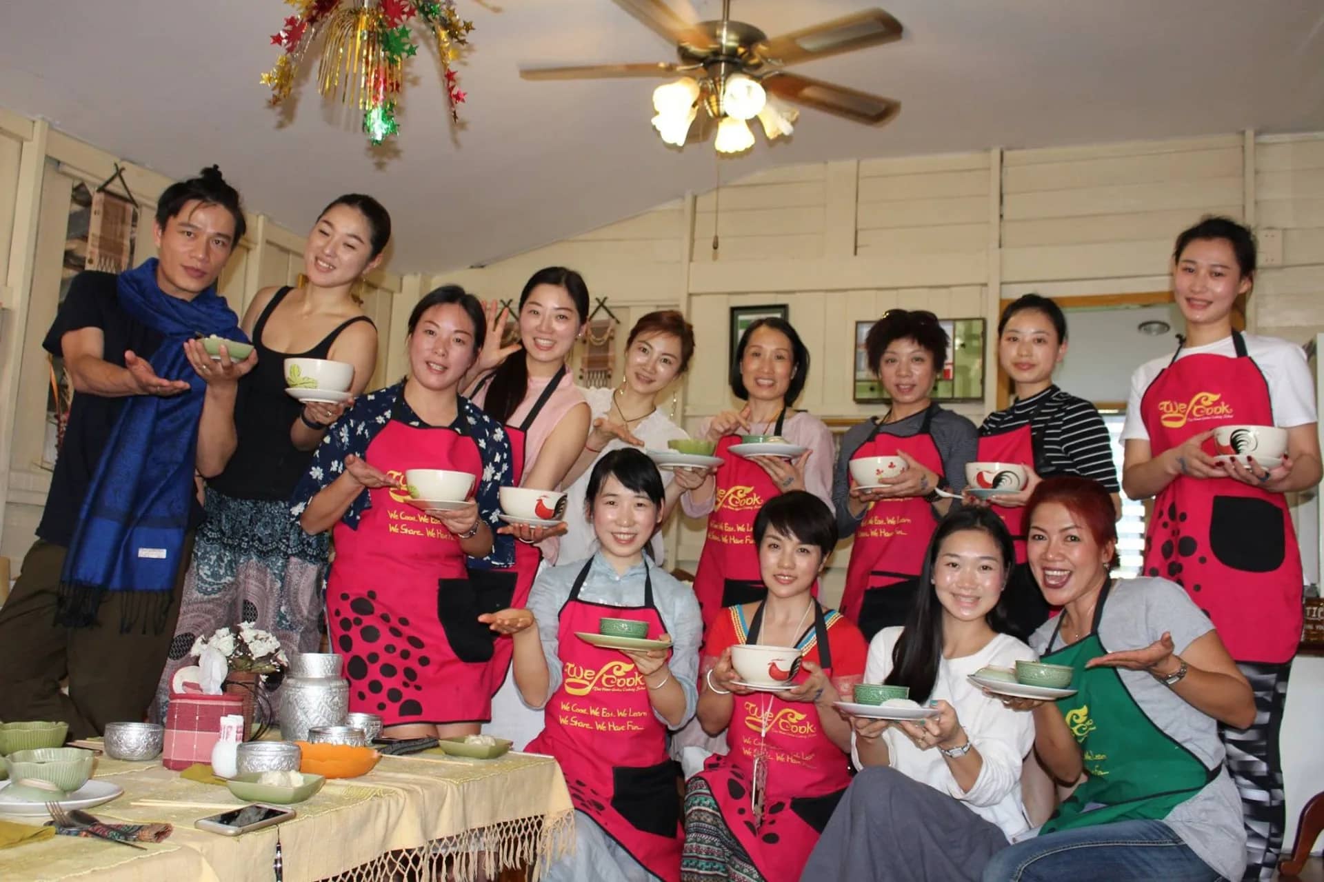 Guests in red aprons hold up bowls of food they cooked at We Cook Thai cooking school