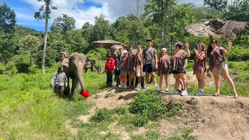 Visitors in matching ponchos pose on a grassy slope while an elephant eats nearby at the sanctuary