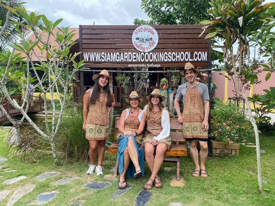 Four guests in straw hats and aprons pose in front of the Siam Garden Cooking School sign