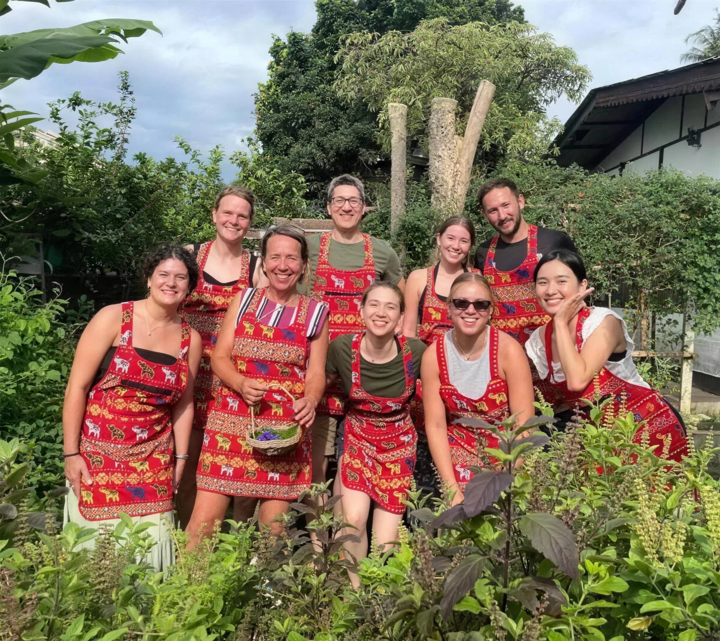 Cooking class guests in red patterned aprons pose in the garden of Thai Cottage Home Cookery School