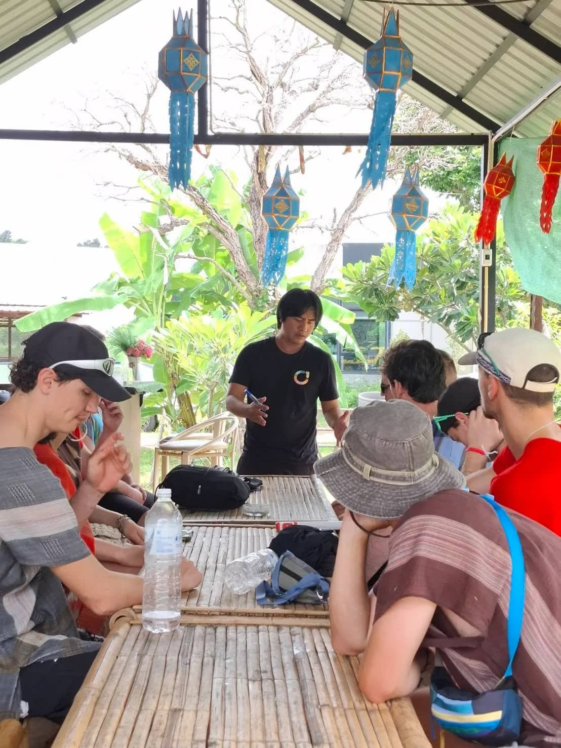 Guests in hats sit at a long bamboo table under a shelter hung with paper lanterns