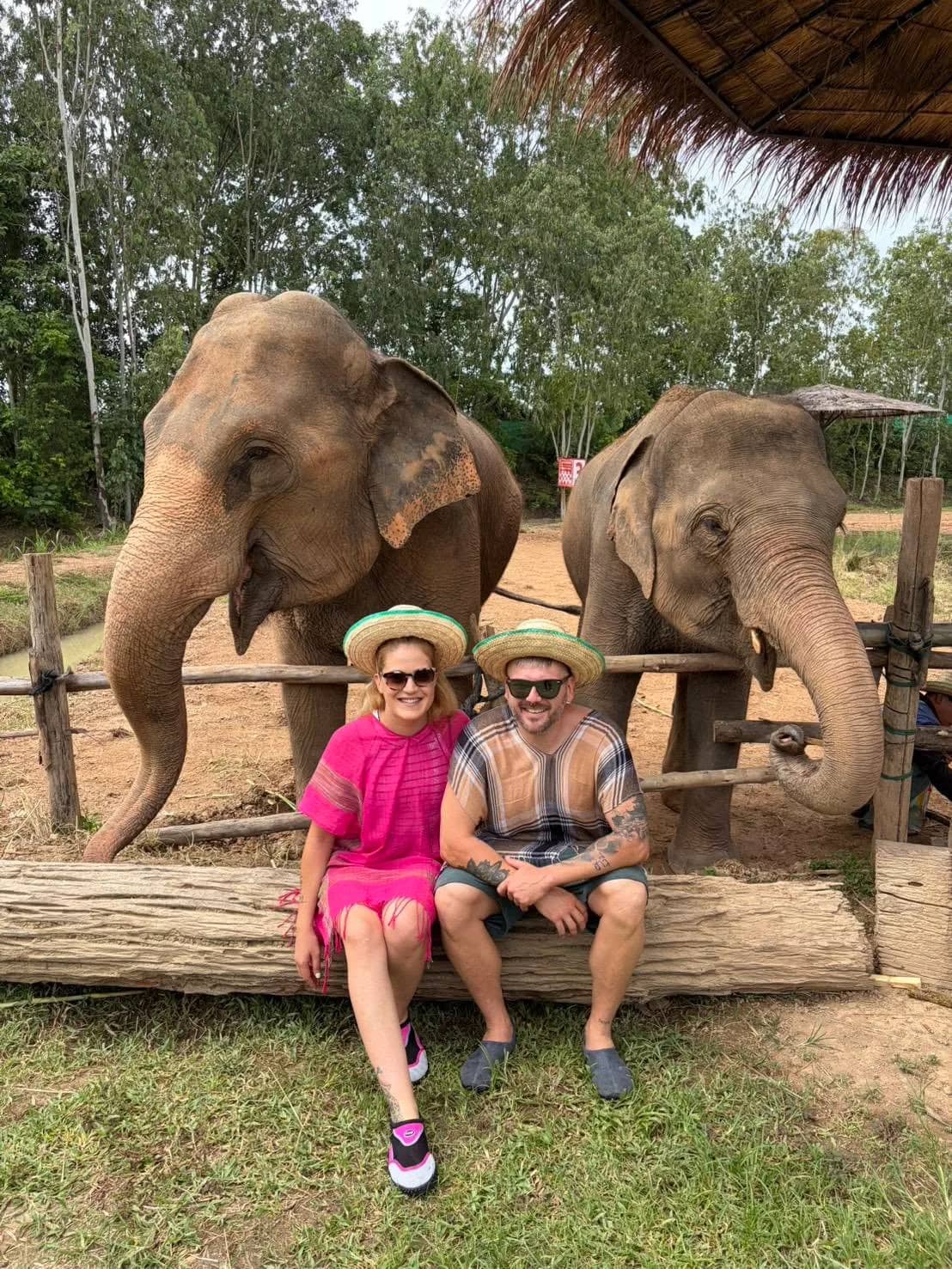 A couple in straw hats sits on a log in front of two elephants behind a wooden rail.