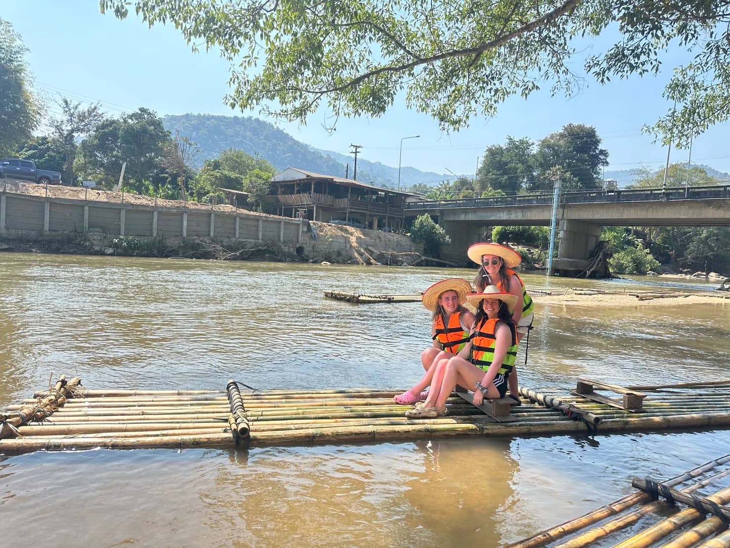 Two guests in straw hats and life jackets sit on a bamboo raft below a river bridge