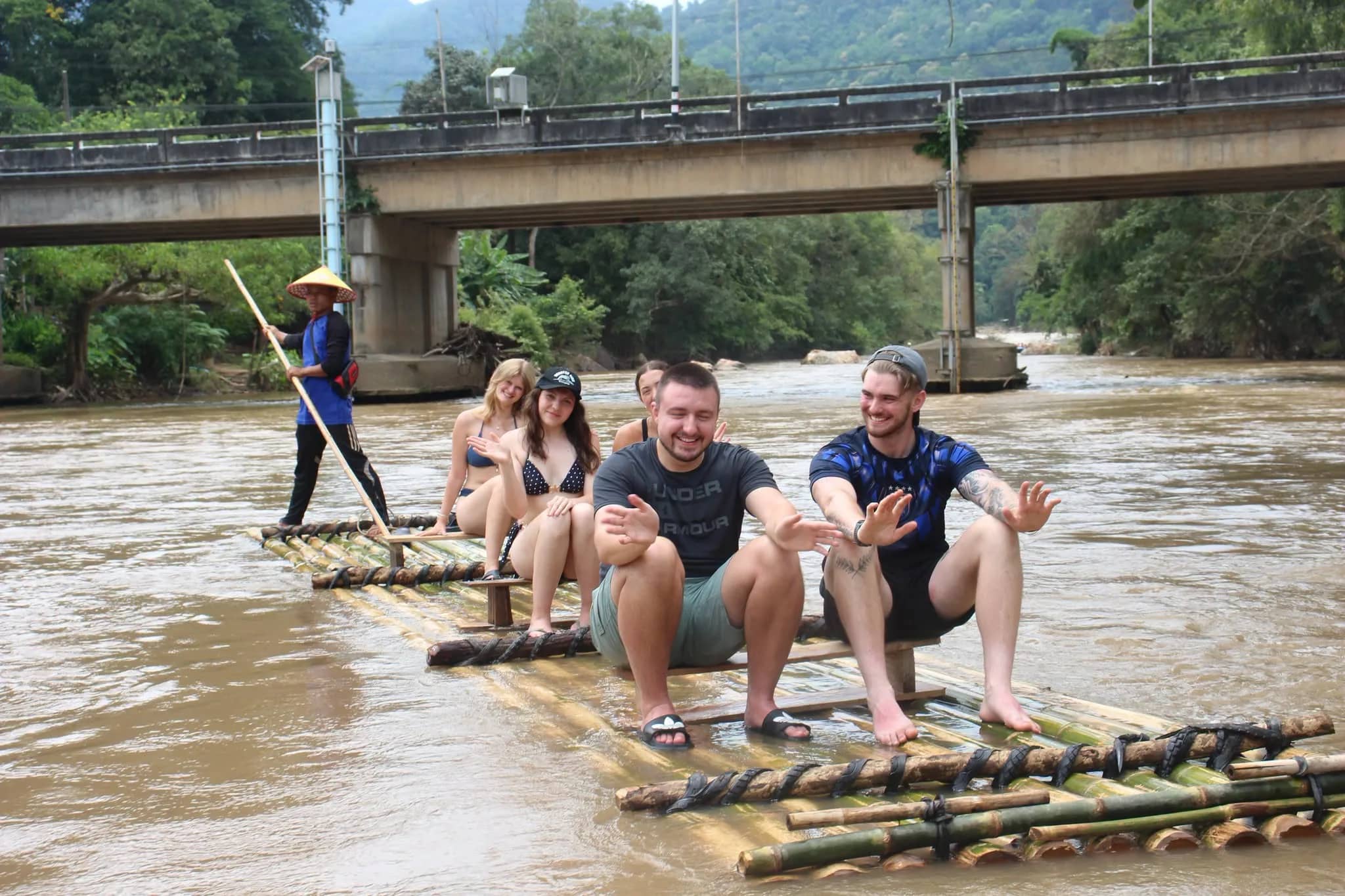 A guide poles a bamboo raft carrying smiling guests down a river beneath a concrete bridge