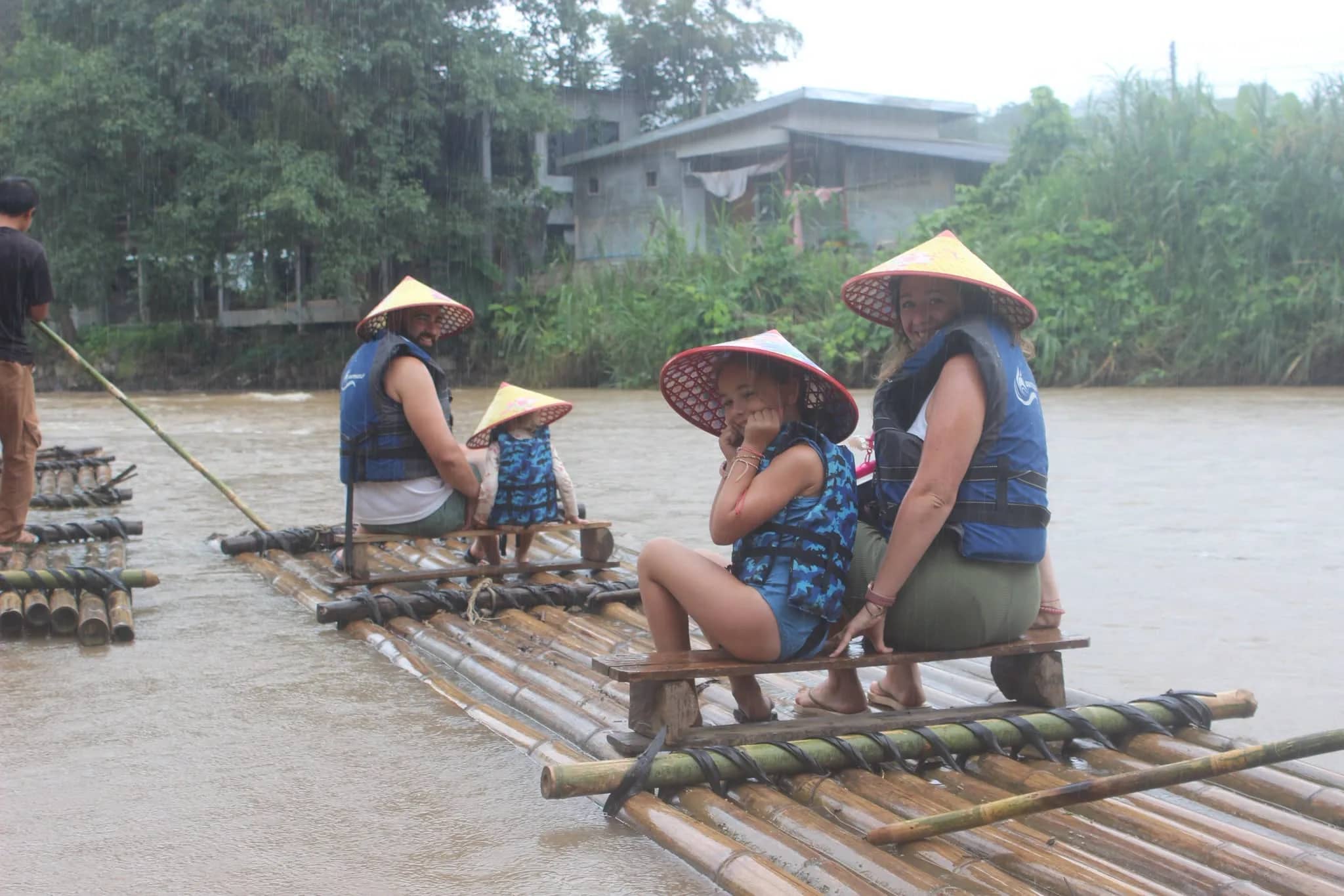 A family in conical straw hats and life jackets sits on a bamboo raft on a misty river