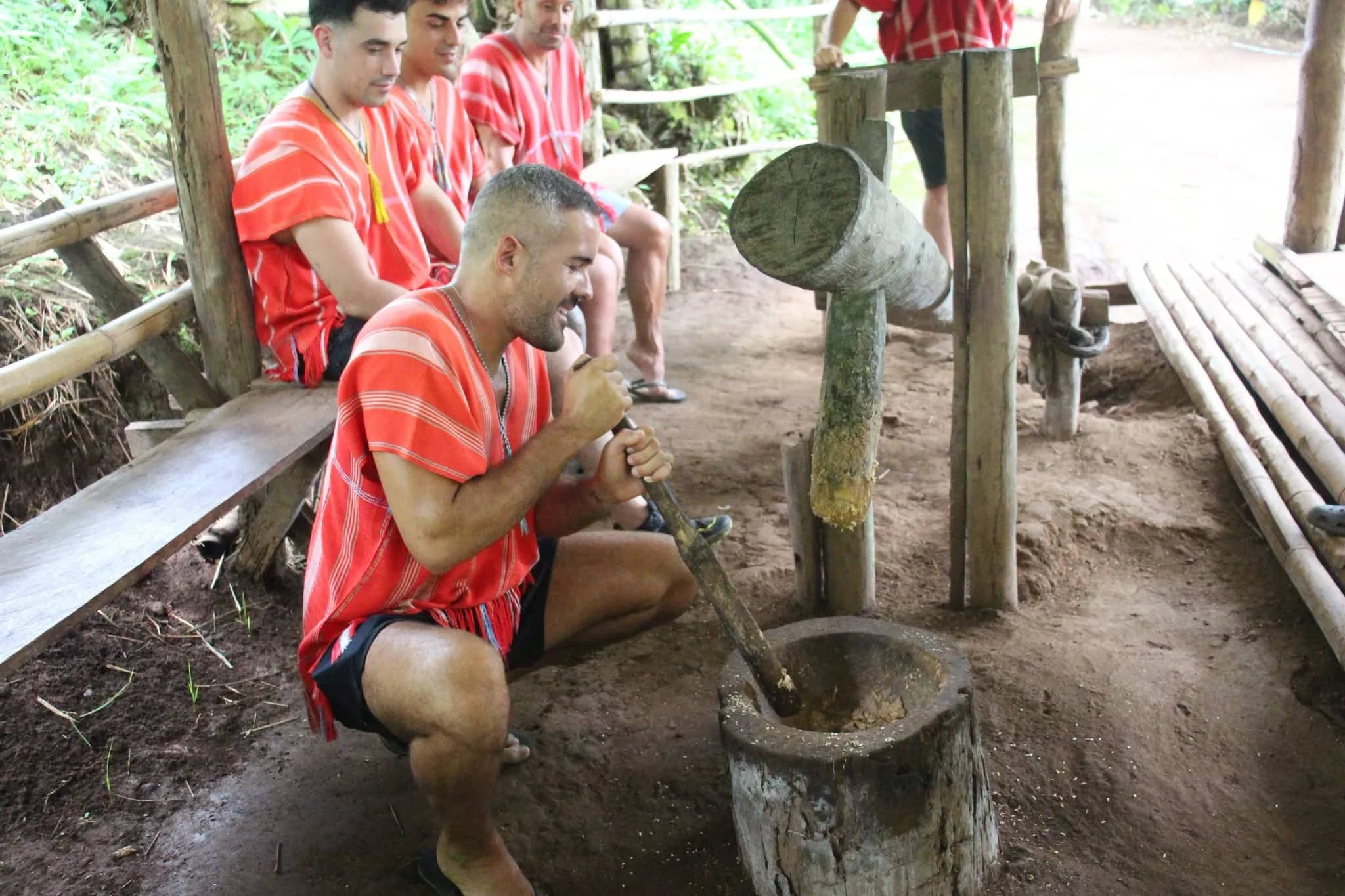 Visitors in red tunics take turns pounding herbs with a long pestle in a wooden mortar