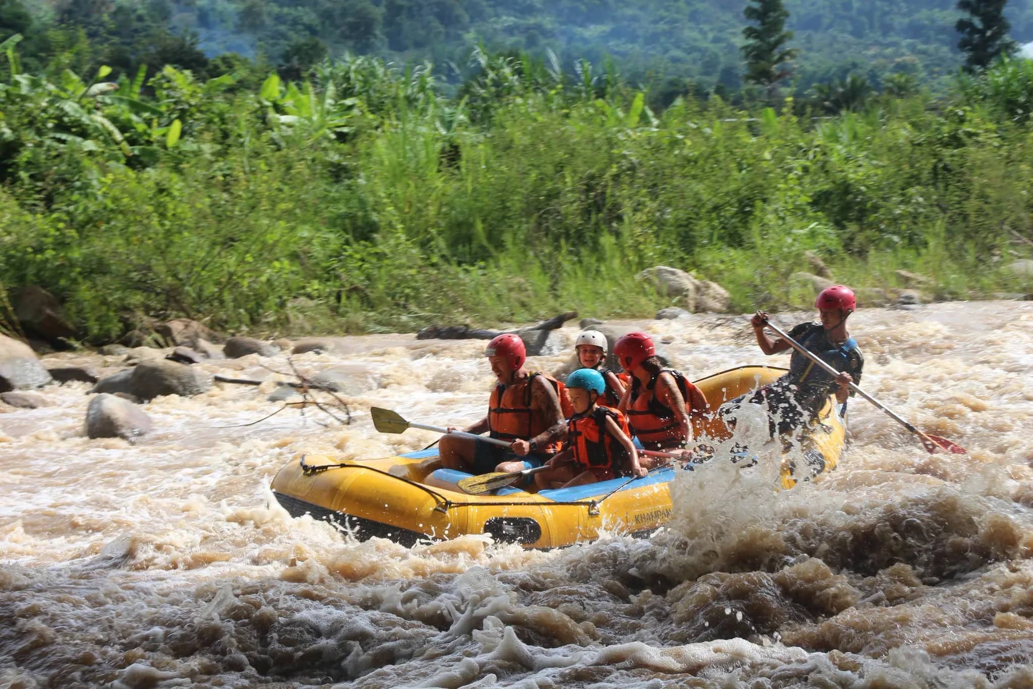 Rafters in red helmets paddle a yellow raft through rushing water on a jungle river