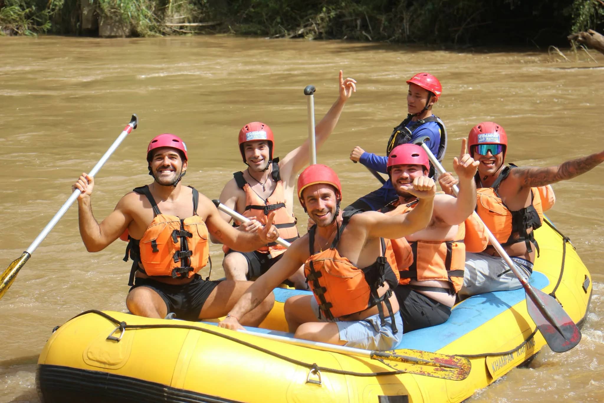 A cheerful crew in red helmets and orange life jackets raise their paddles in a yellow raft
