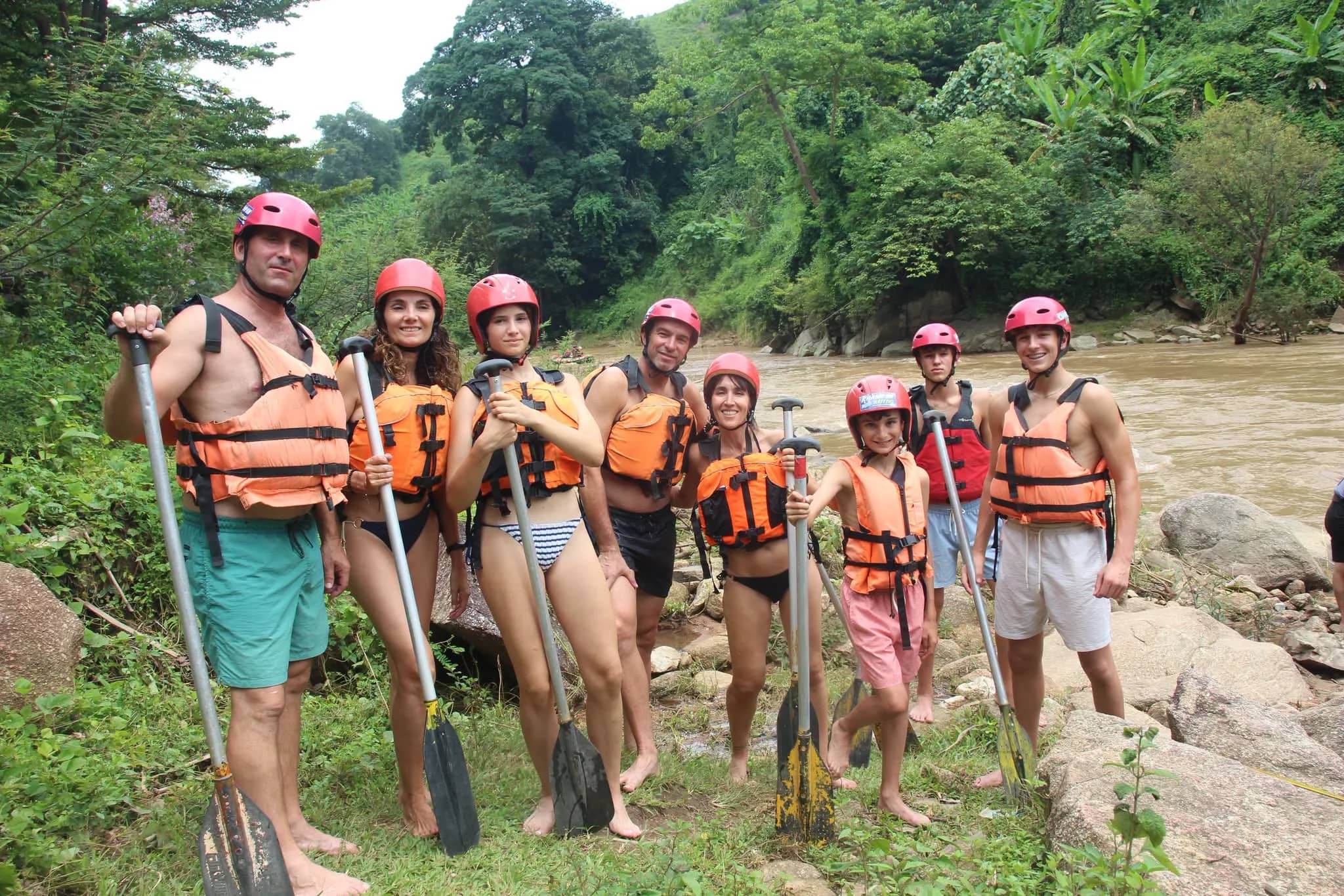 A group in red helmets and orange life jackets stands with paddles on the rocky river bank