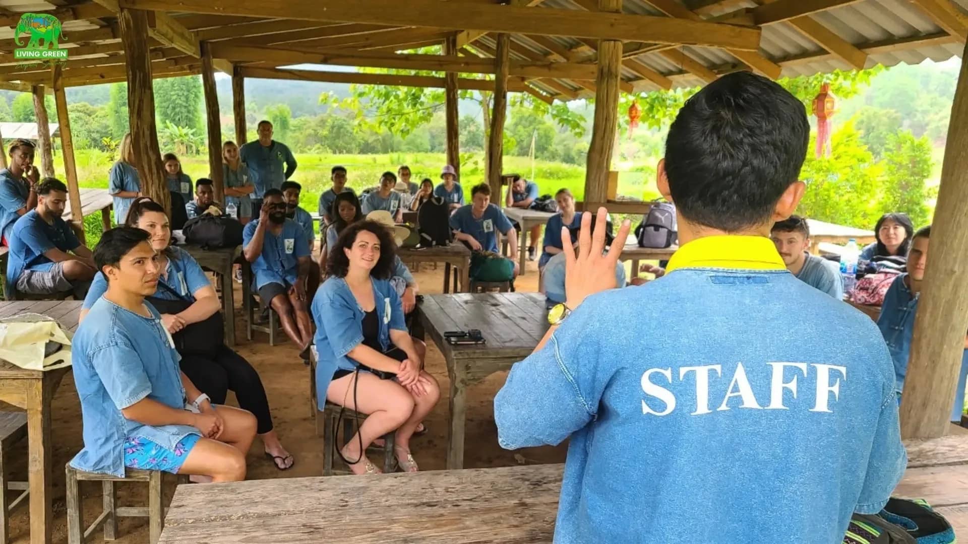 A staff member gives a welcome talk to visitors seated at tables in an open-air sala