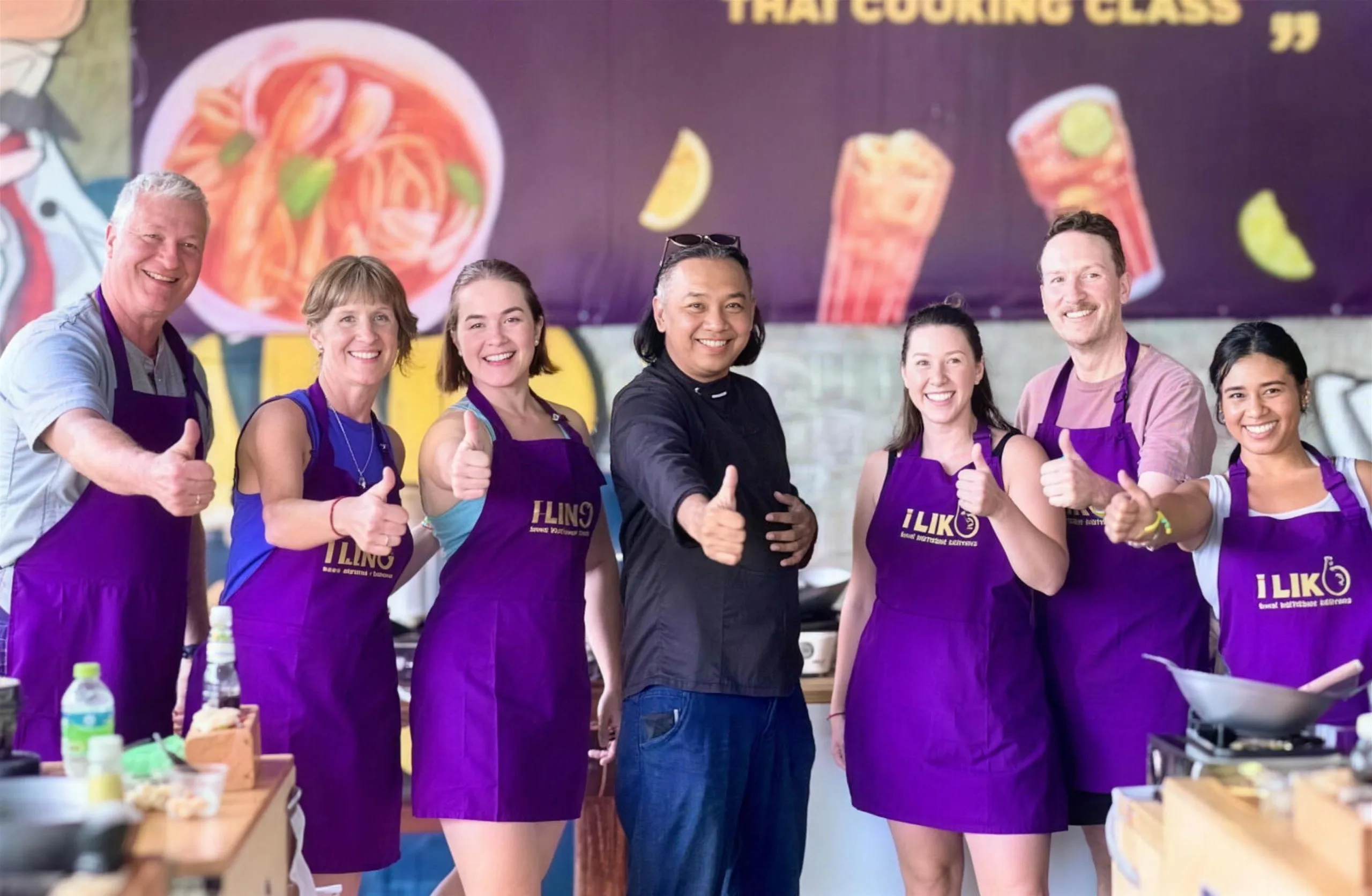 Cooking class guests in purple aprons give a thumbs up with their chef at i-Like Thai Cooking School