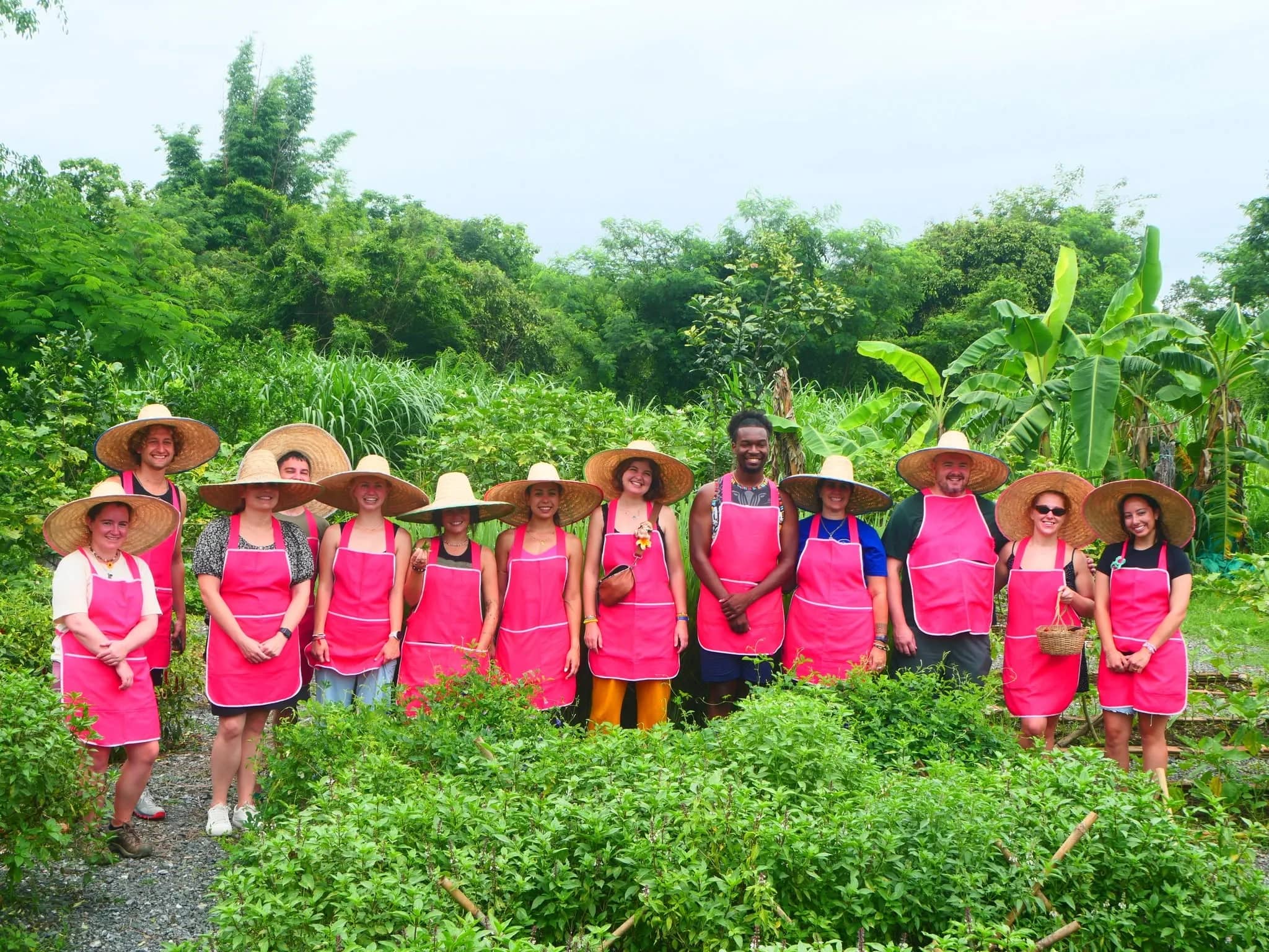 Guests in pink aprons and straw hats stand among vegetables at Smile Organic Farm Cooking School