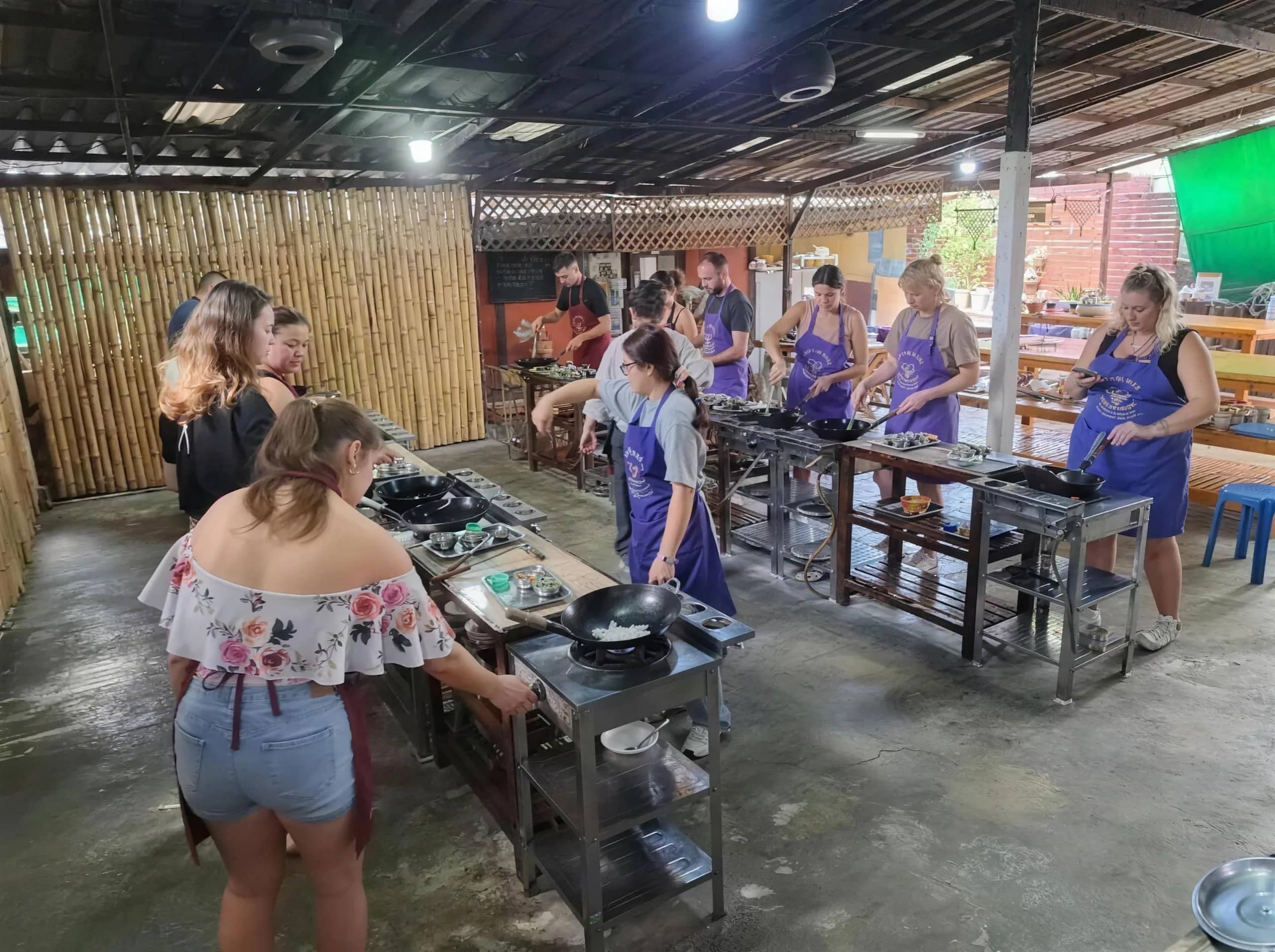 Guests in aprons cook at individual wok stations in the open kitchen of a Thai cooking school