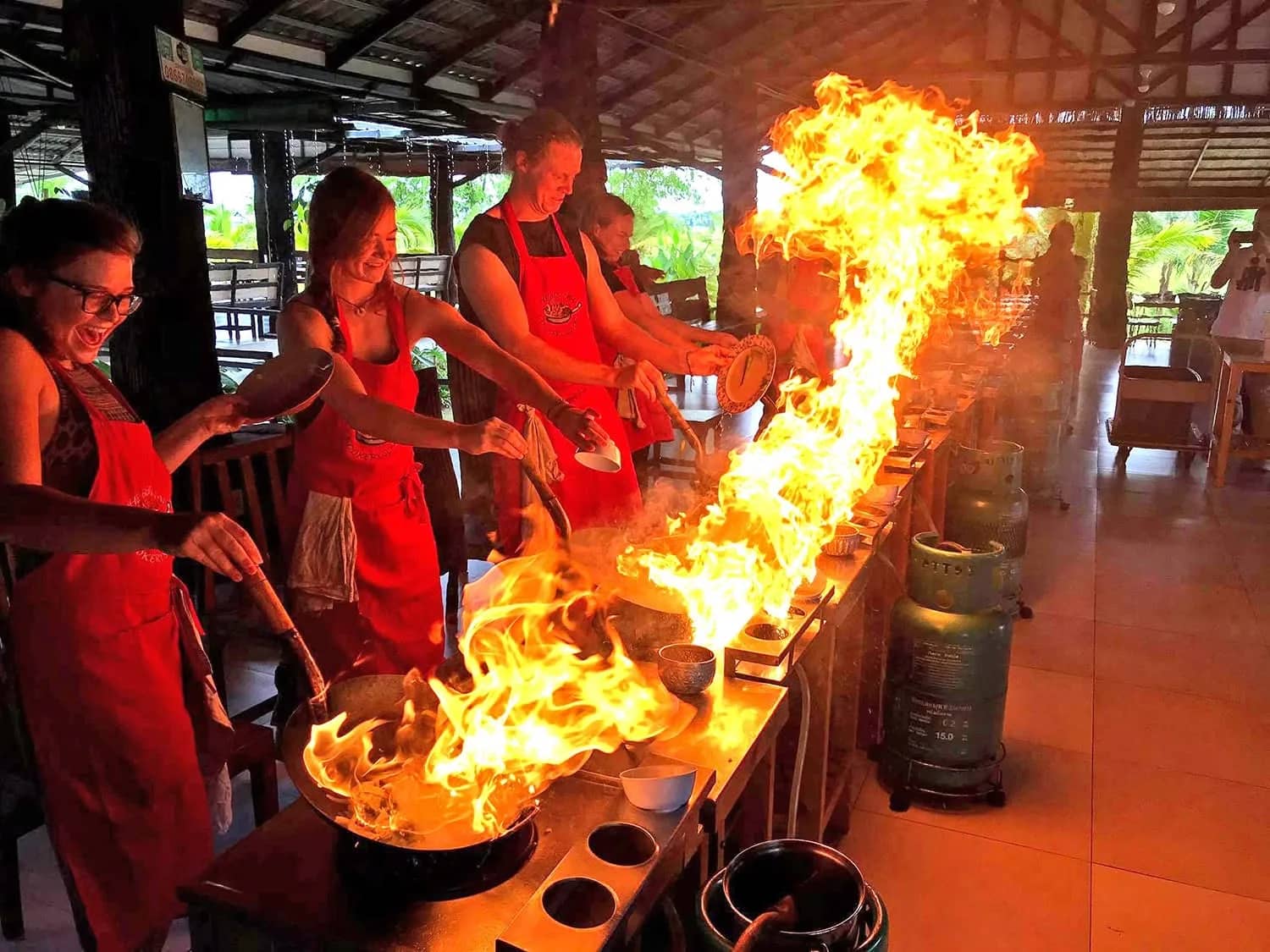 Guests in red aprons cook as tall flames burst from a wok at an open-air cooking school