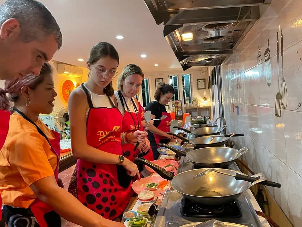 Guests in red aprons cook at a row of woks along a tiled kitchen counter