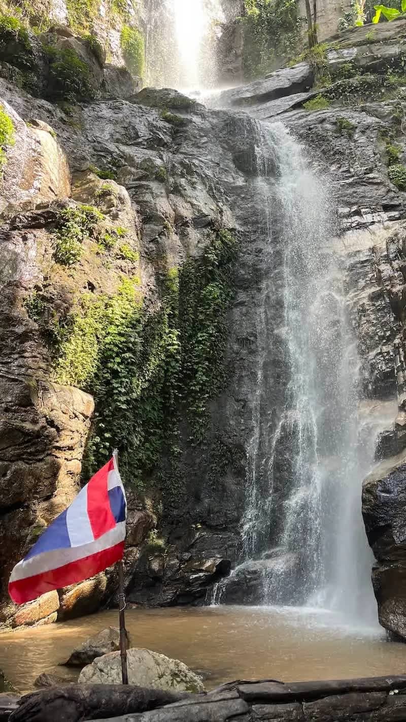 A Thai flag stands at the base of a tall waterfall flowing down a rocky cliff