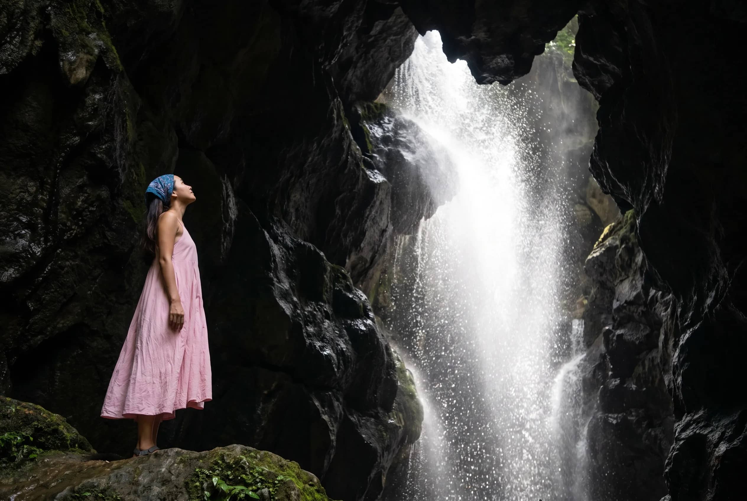A woman in a pink dress stands on a rock ledge looking up at a waterfall pouring into a dark cave