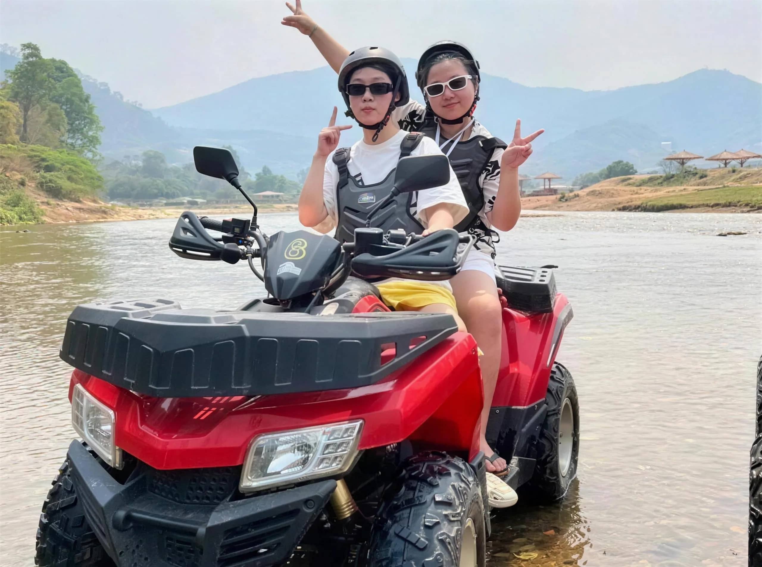 Two riders in helmets wave from a red ATV parked in a shallow river near Chiang Mai