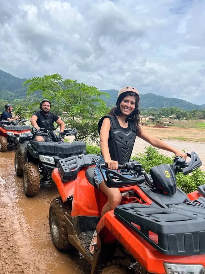 Two smiling riders in helmets sit on red ATV quad bikes on a muddy track by the hills