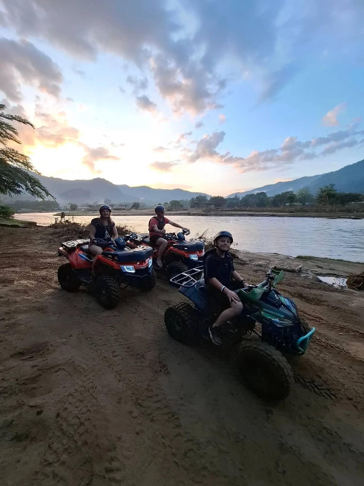 Riders on quad bikes stop on a sandy riverbank under a colourful sunset sky