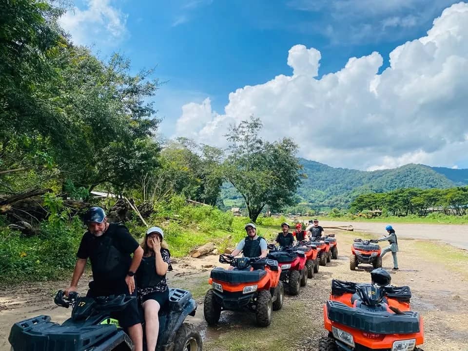 Riders on red and black quad bikes line up on a dirt track with green hills behind them