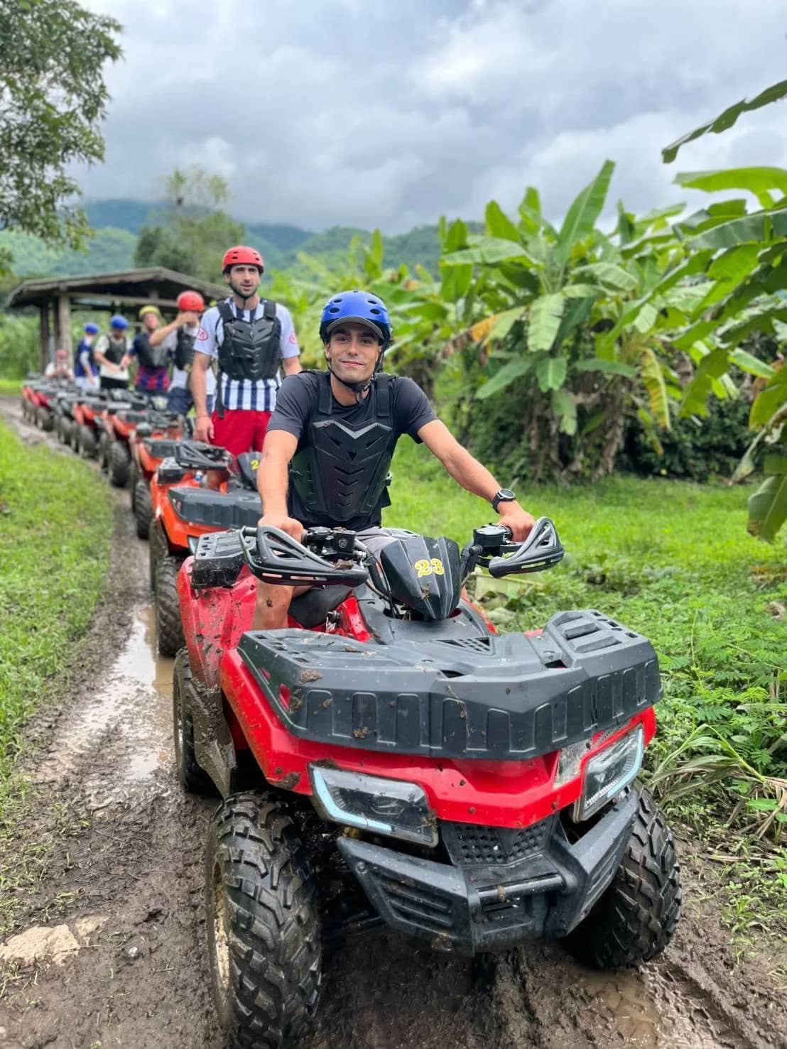 Riders in helmets drive red ATVs in single file along a muddy track past banana plants.