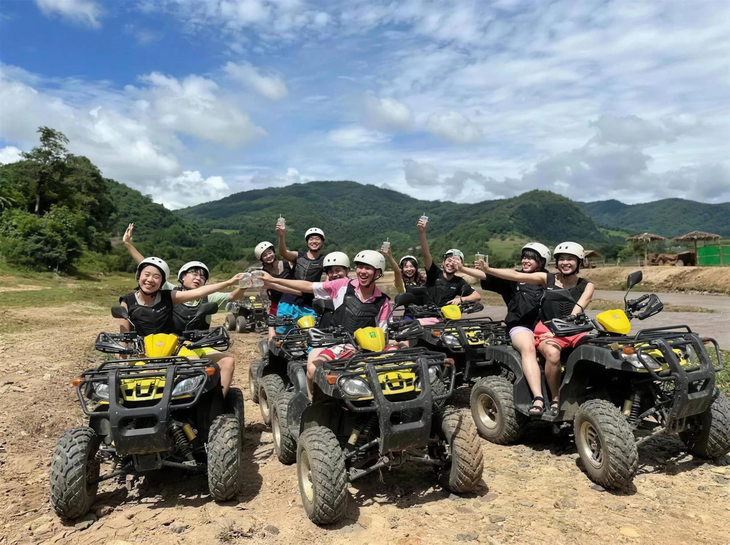 A group of riders in helmets cheers from their ATVs on a dirt field below green mountains