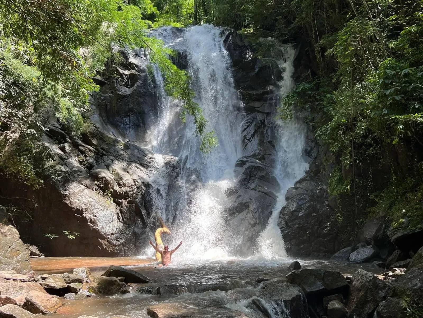 A visitor slides down a tall waterfall into a shallow pool in a forested rock gorge.
