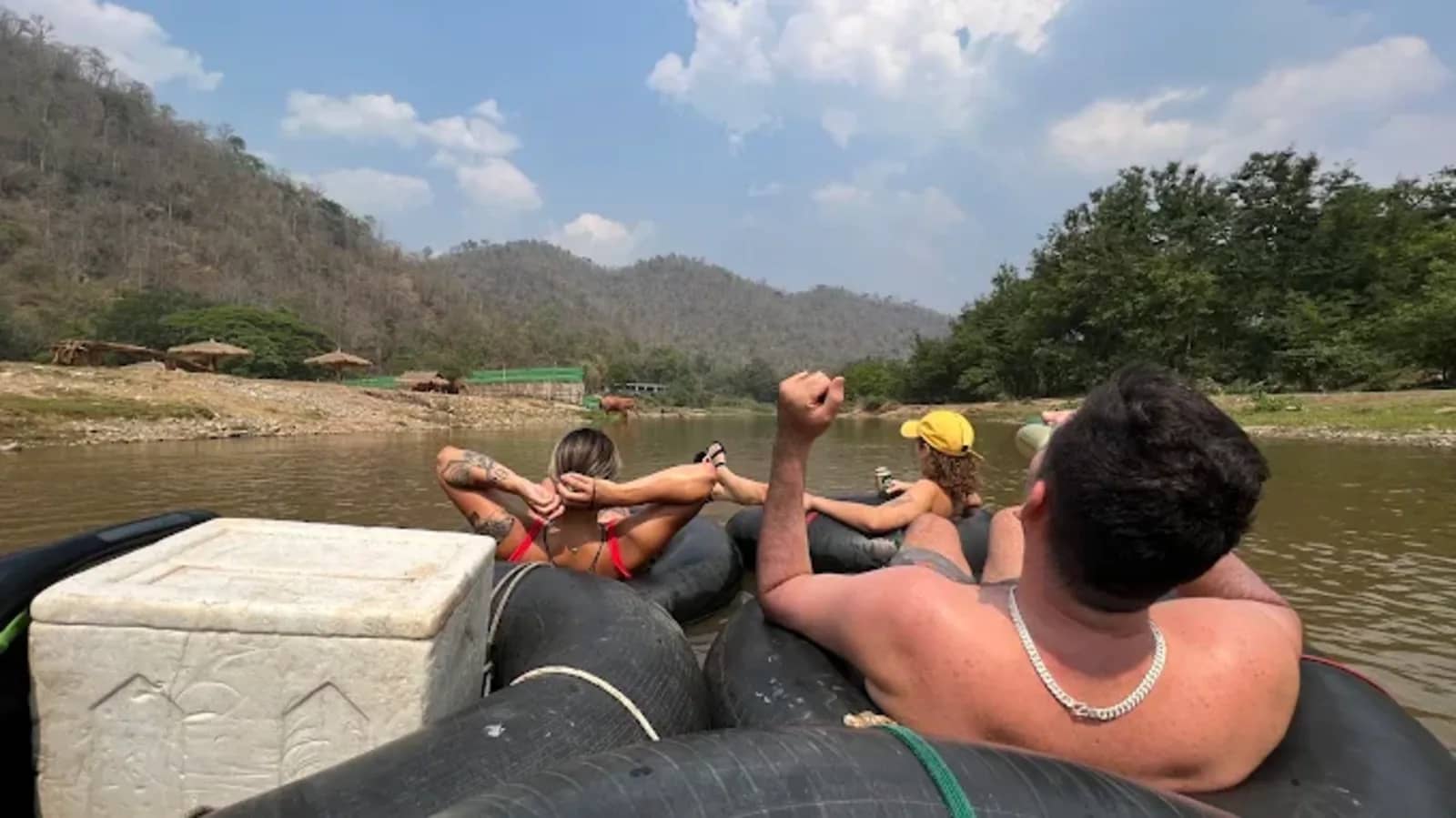 Guests lie back in linked rubber tubes beside a cooler box, drifting along a wide river under blue sky.