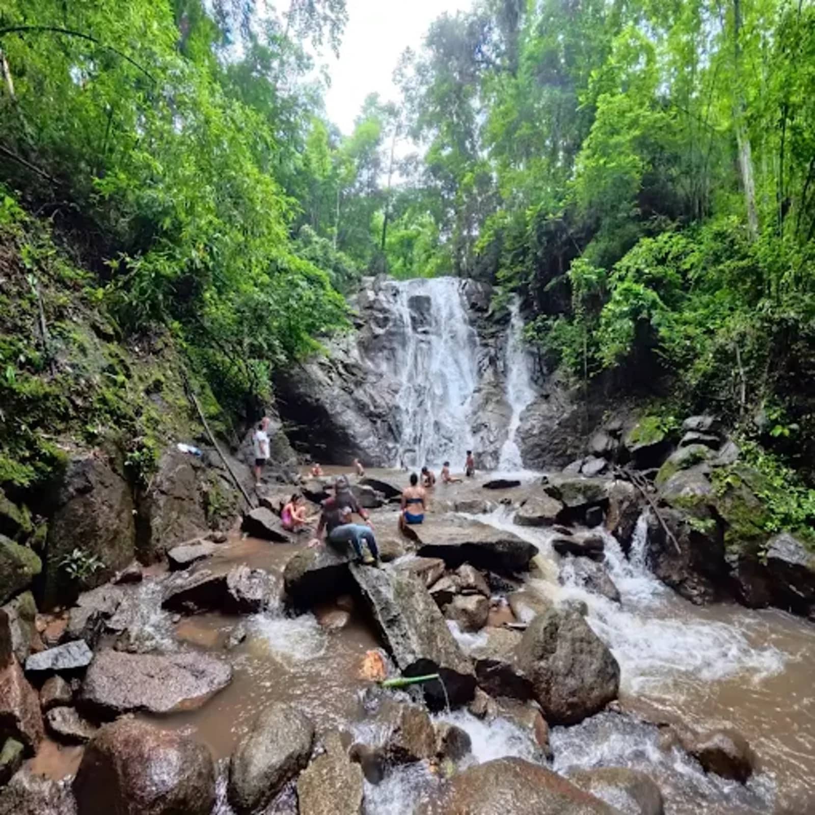Visitors clamber over boulders at the base of a cascading waterfall surrounded by jungle.