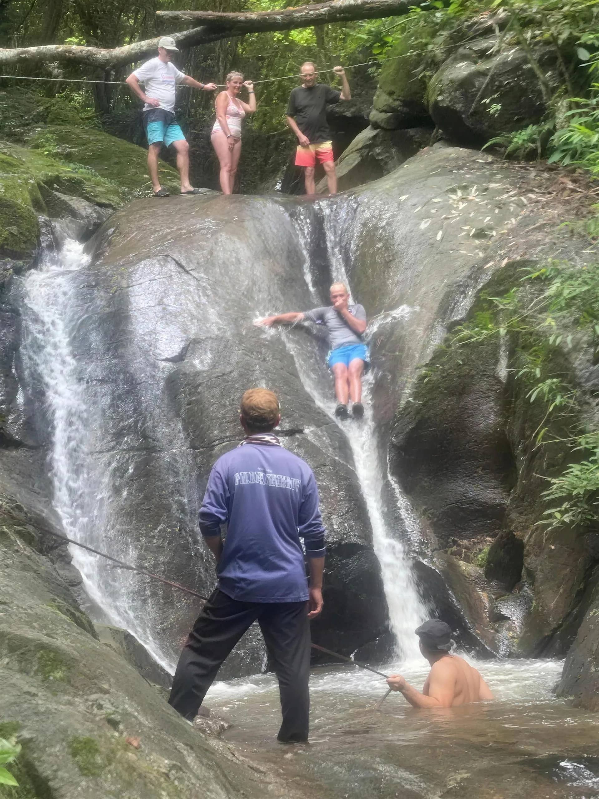 A visitor slides down a smooth rock waterfall while a guide watches from the pool below.