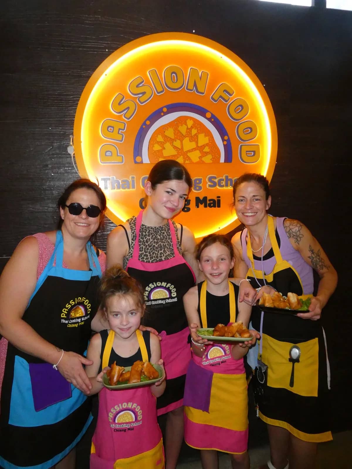 A family in aprons holds plates of food in front of the glowing Passion Food school sign