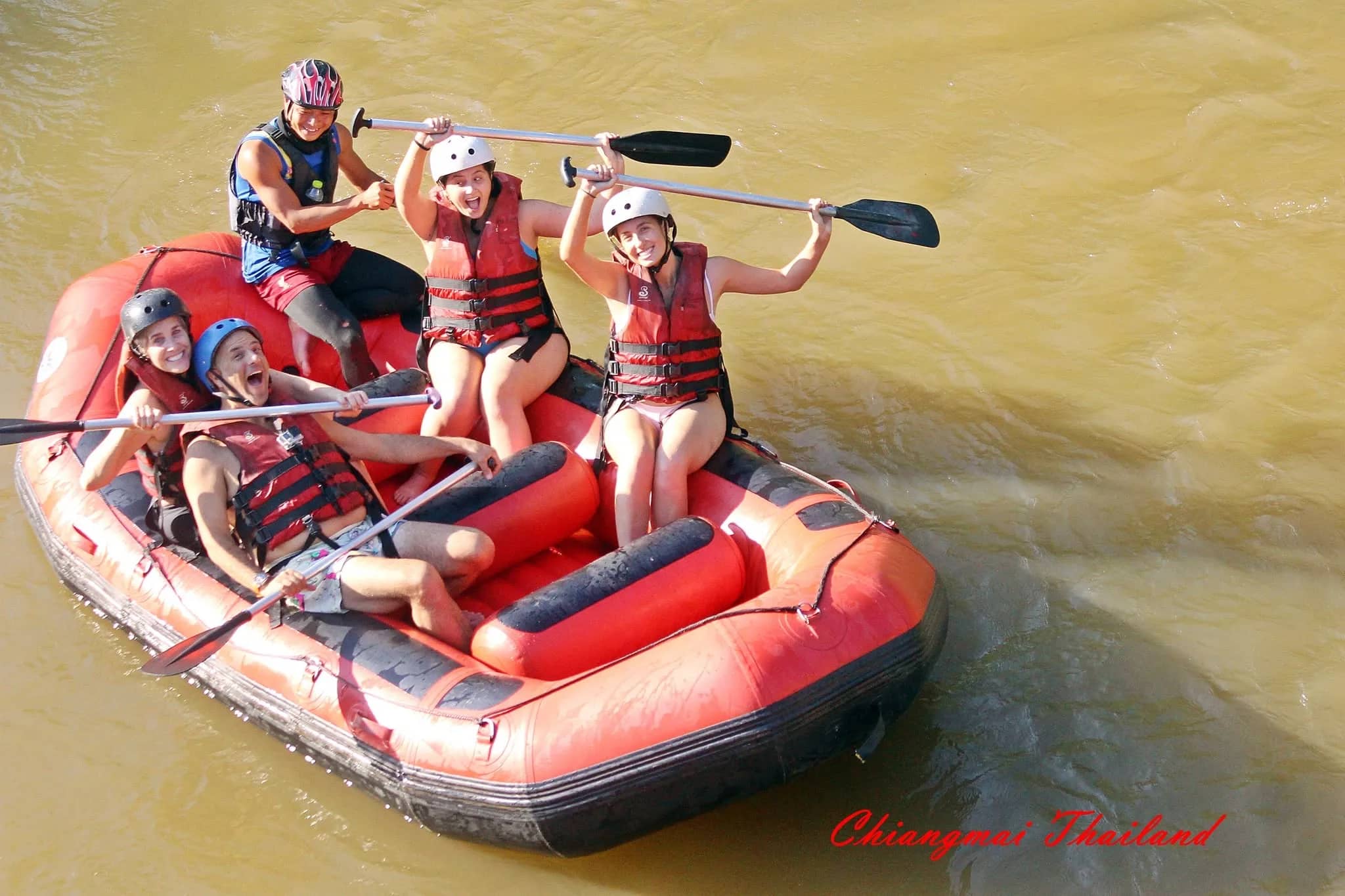 Smiling rafters in helmets and life jackets raise their paddles in a red raft on a brown river.