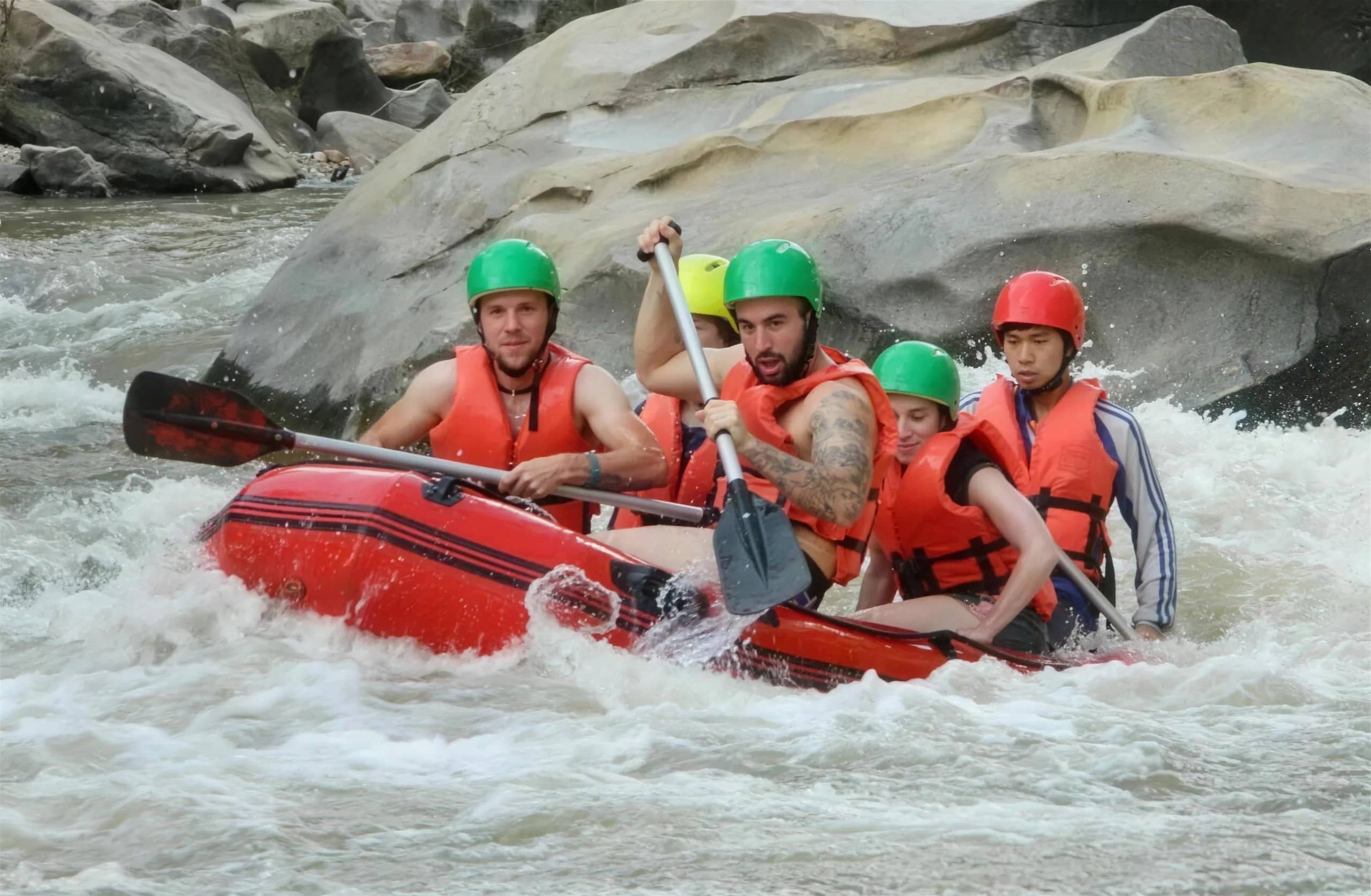 Rafters in green and red helmets paddle a red raft through foaming rapids beside a large boulder.