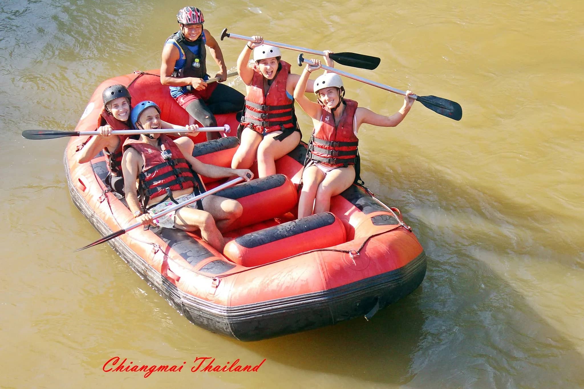 Rafters in helmets and life jackets lift their paddles in a red raft on a brown river