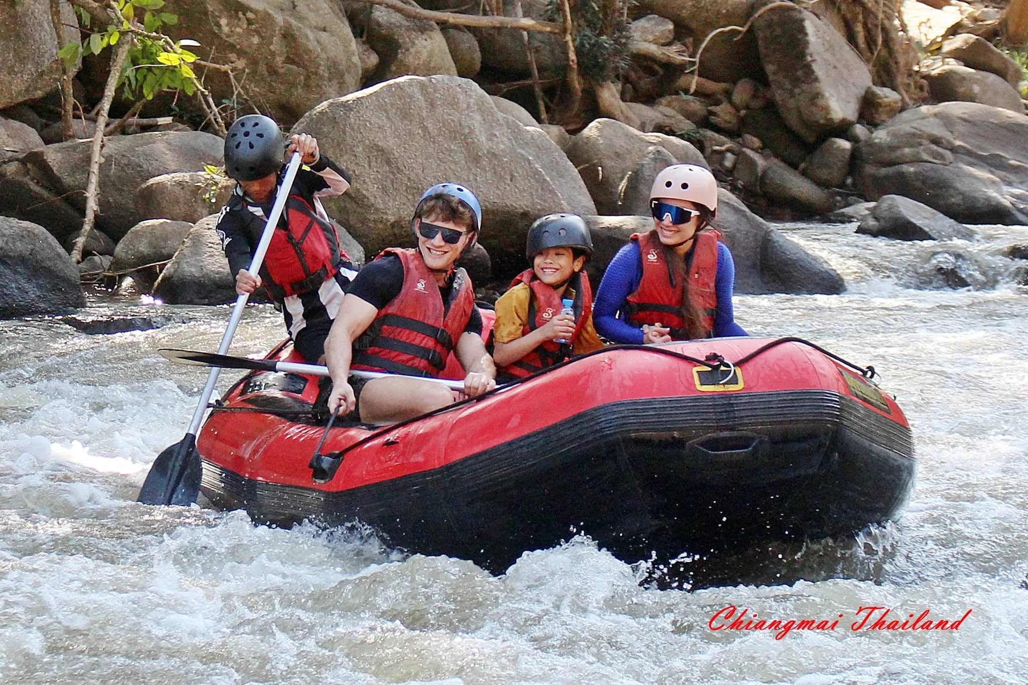 Four rafters in helmets and life jackets ride a red raft past large rocks in fast white water.