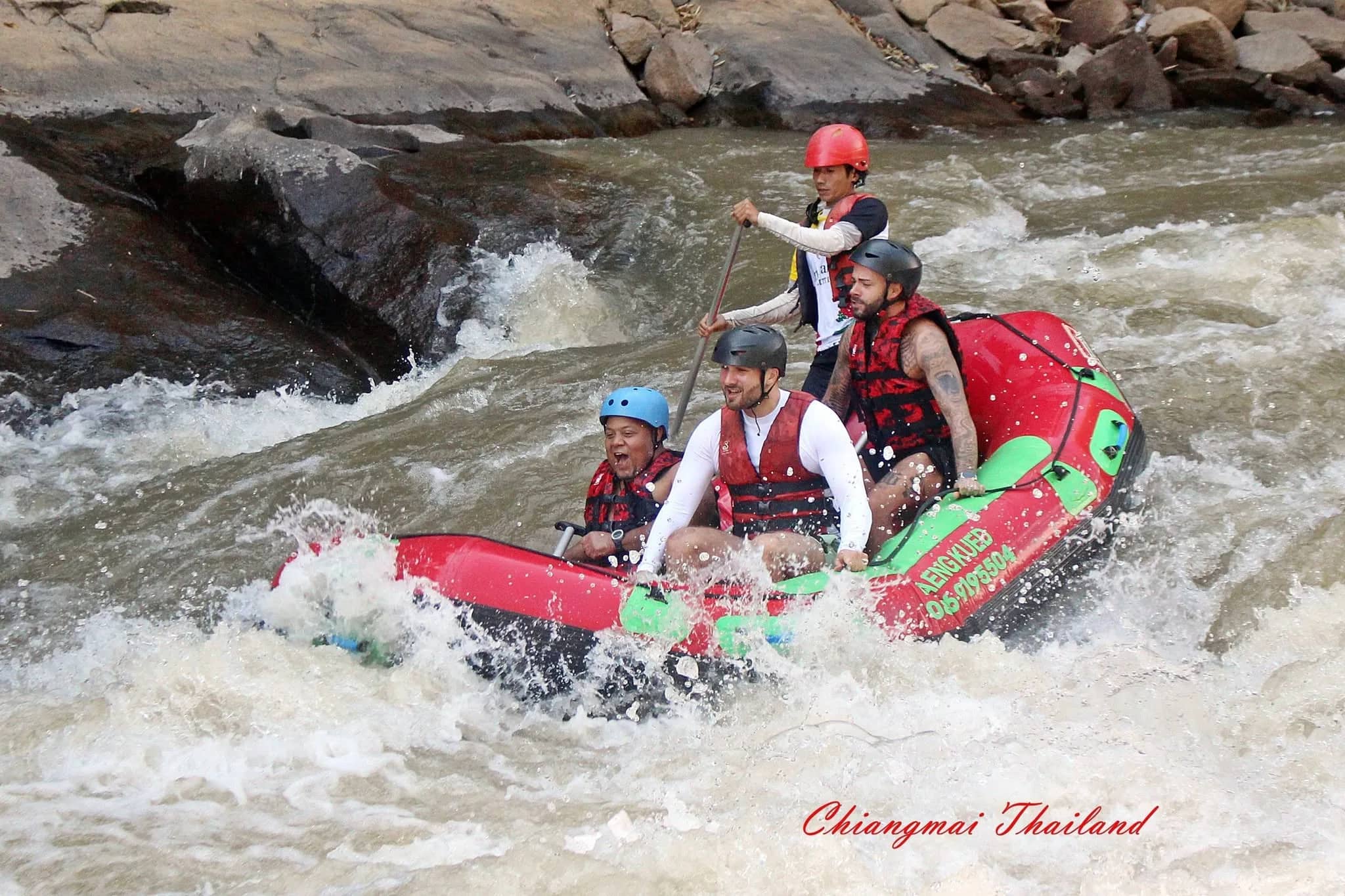 A crew in helmets and life jackets paddles a red and green raft through white rapids near Chiang Mai