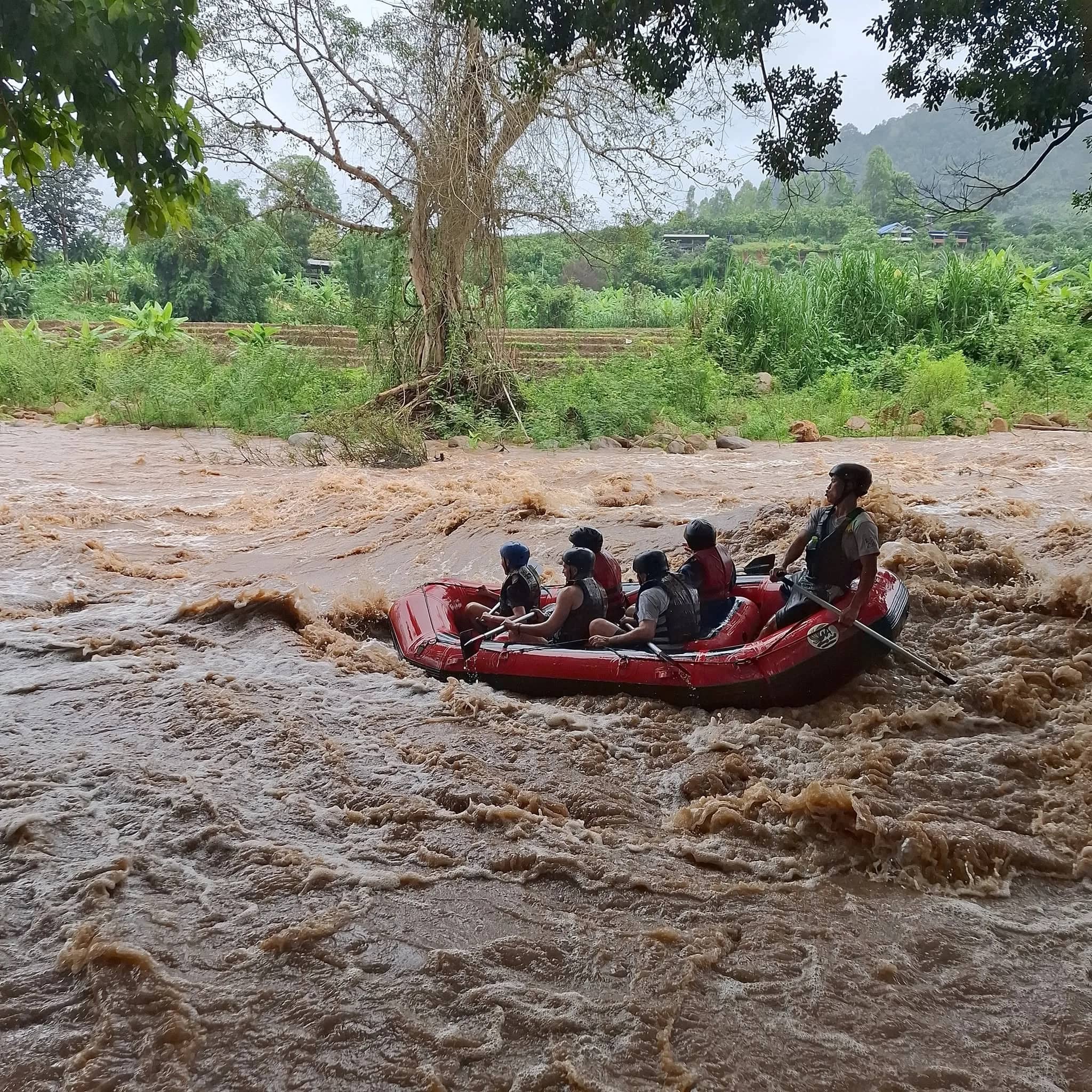 Rafters in life jackets paddle a red raft across churning brown water beside green fields