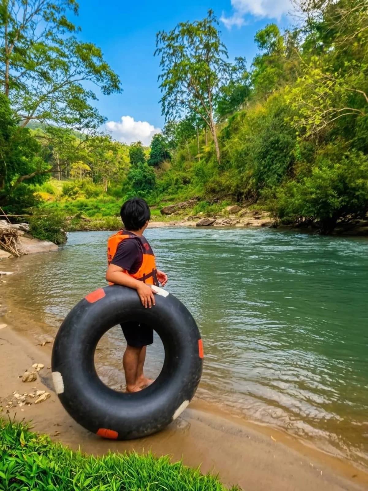 A guest in a life jacket holds a large black tube on the bank of a clear jungle river