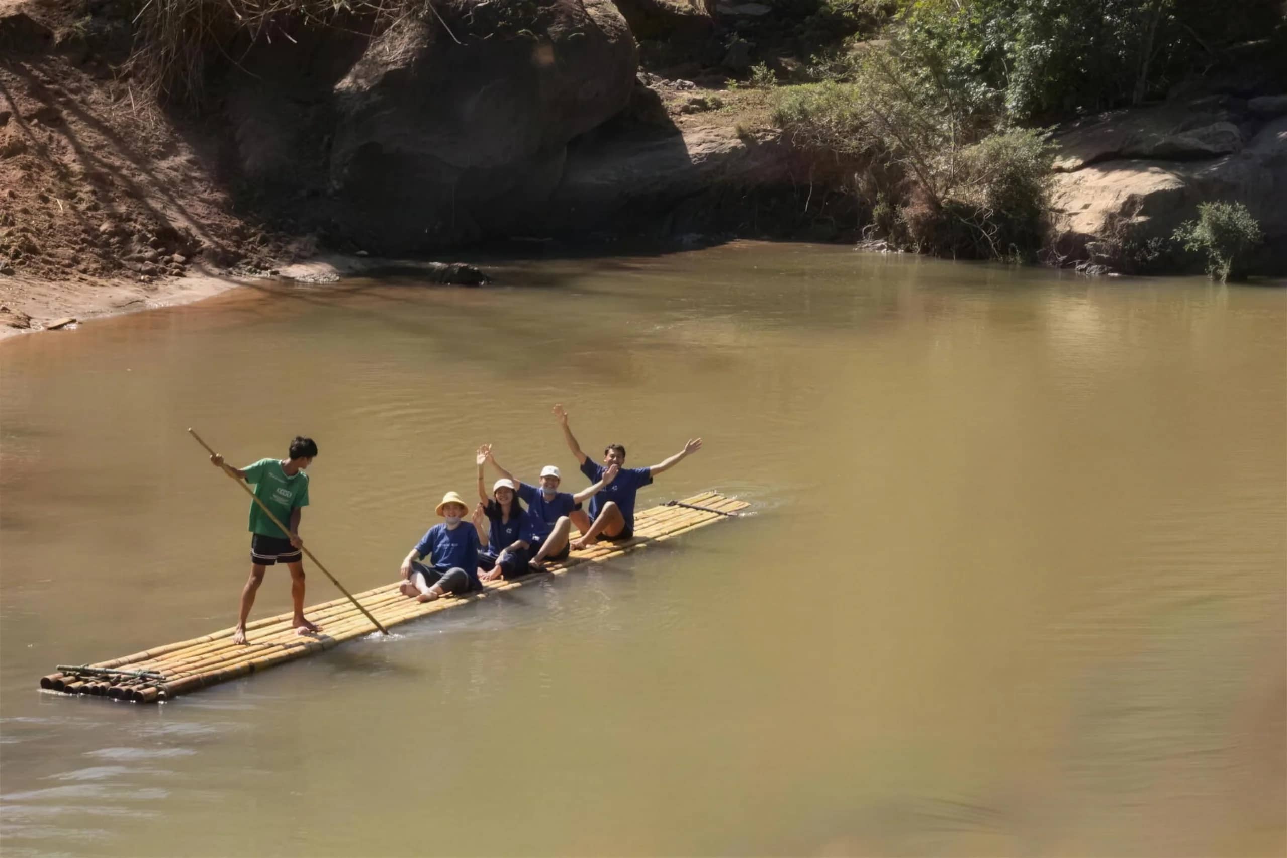 Visitors cheer with raised arms on a bamboo raft steered by a guide with a long pole