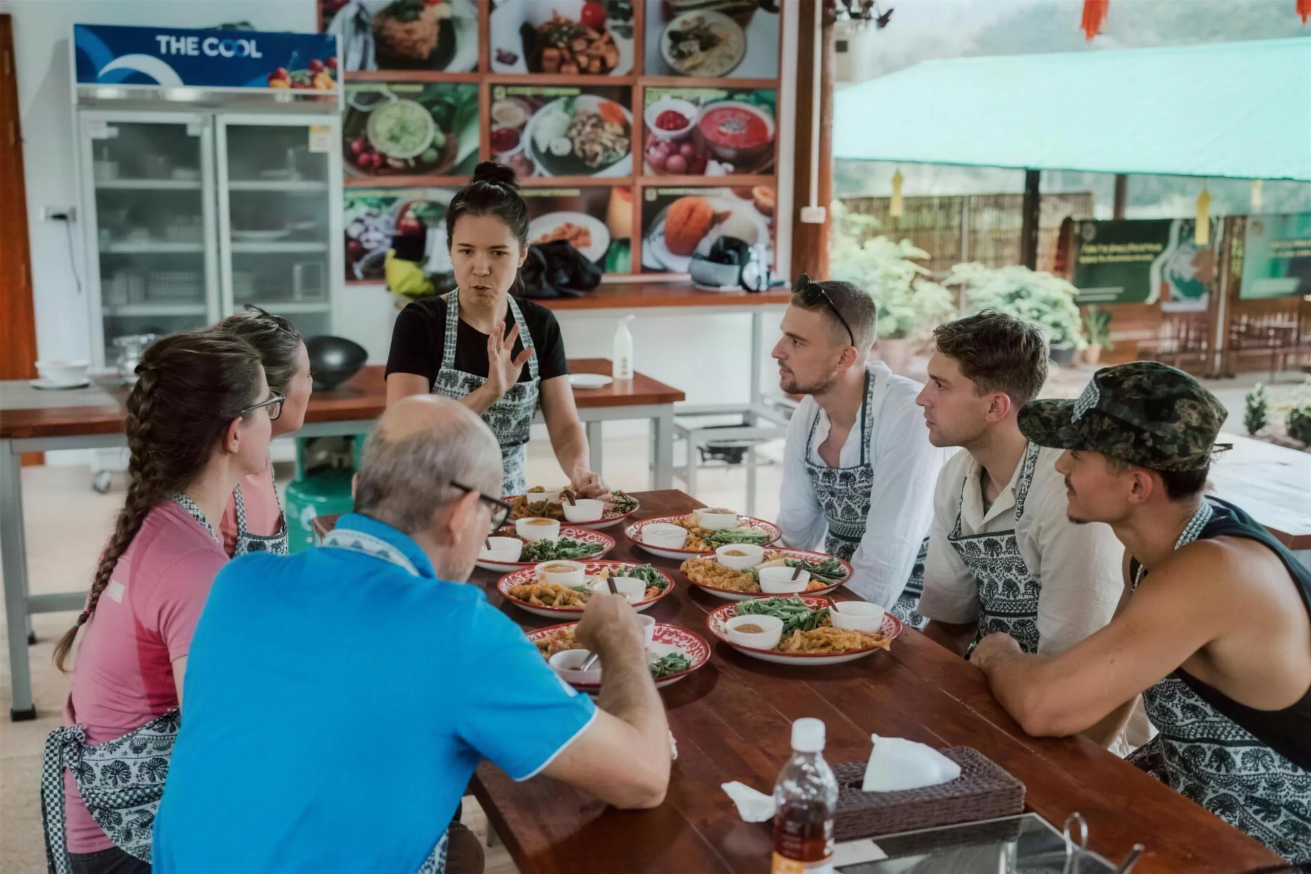 An instructor talks with guests seated at a table of freshly cooked Thai dishes in an open kitchen.
