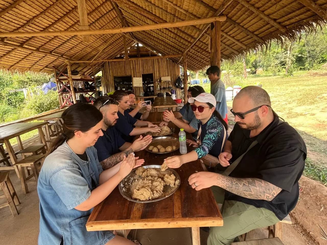 Guests mash food together in metal bowls at a long wooden table under a thatched roof
