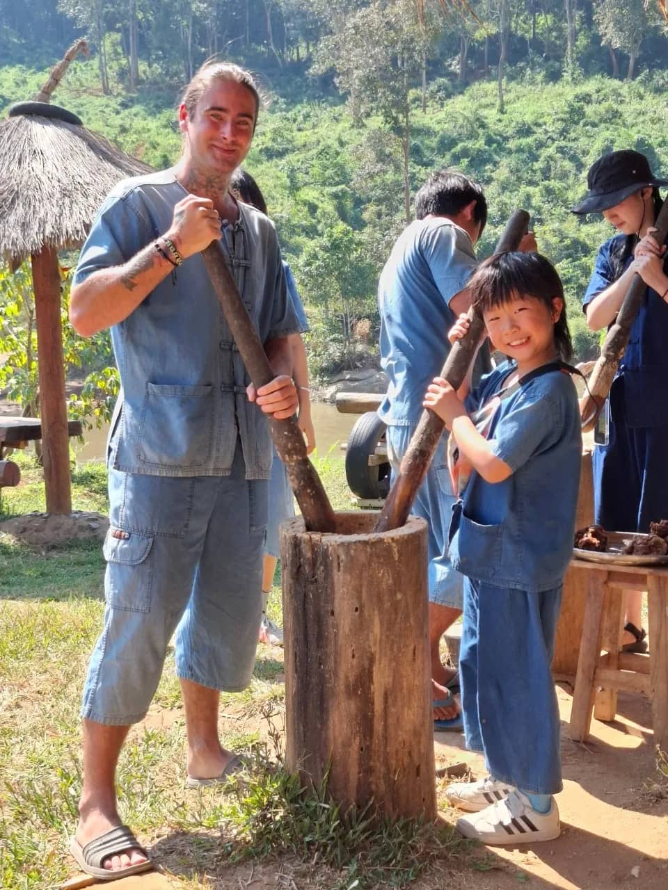 A visitor and a child pound rice with long wooden pestles in a traditional mortar at the sanctuary.