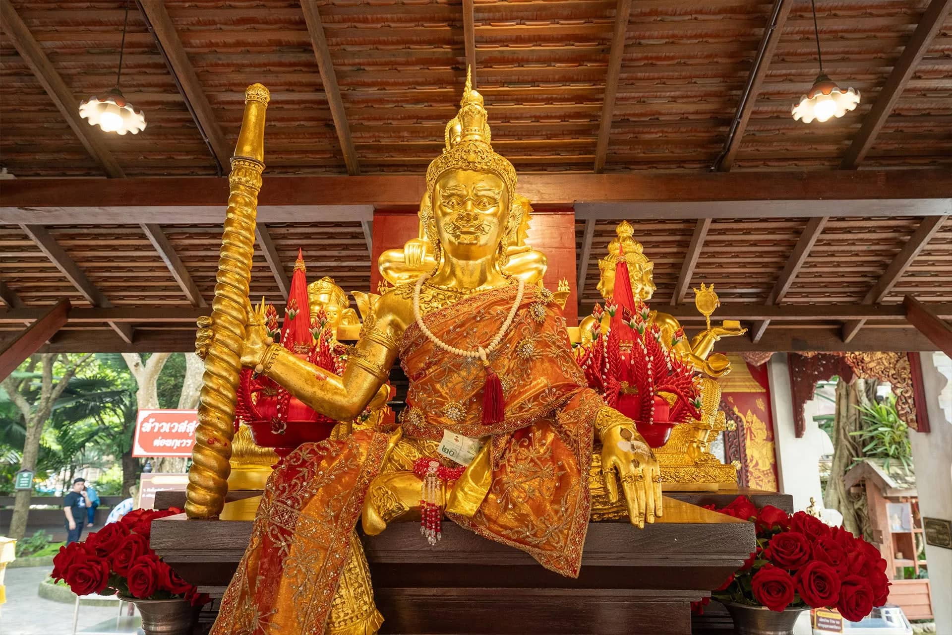 A golden seated deity statue with an ornate robe sits under a wooden pavilion with red roses